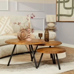 De Salontafel 70 x 70 CM, een ronde houten salontafel met zwart metalen poten, is gestyled in een woonkamer met een roze kandelaar, glazen vaas en keramische kan. Een beige bank en moderne kunstwerken maken de achtergrond compleet.
