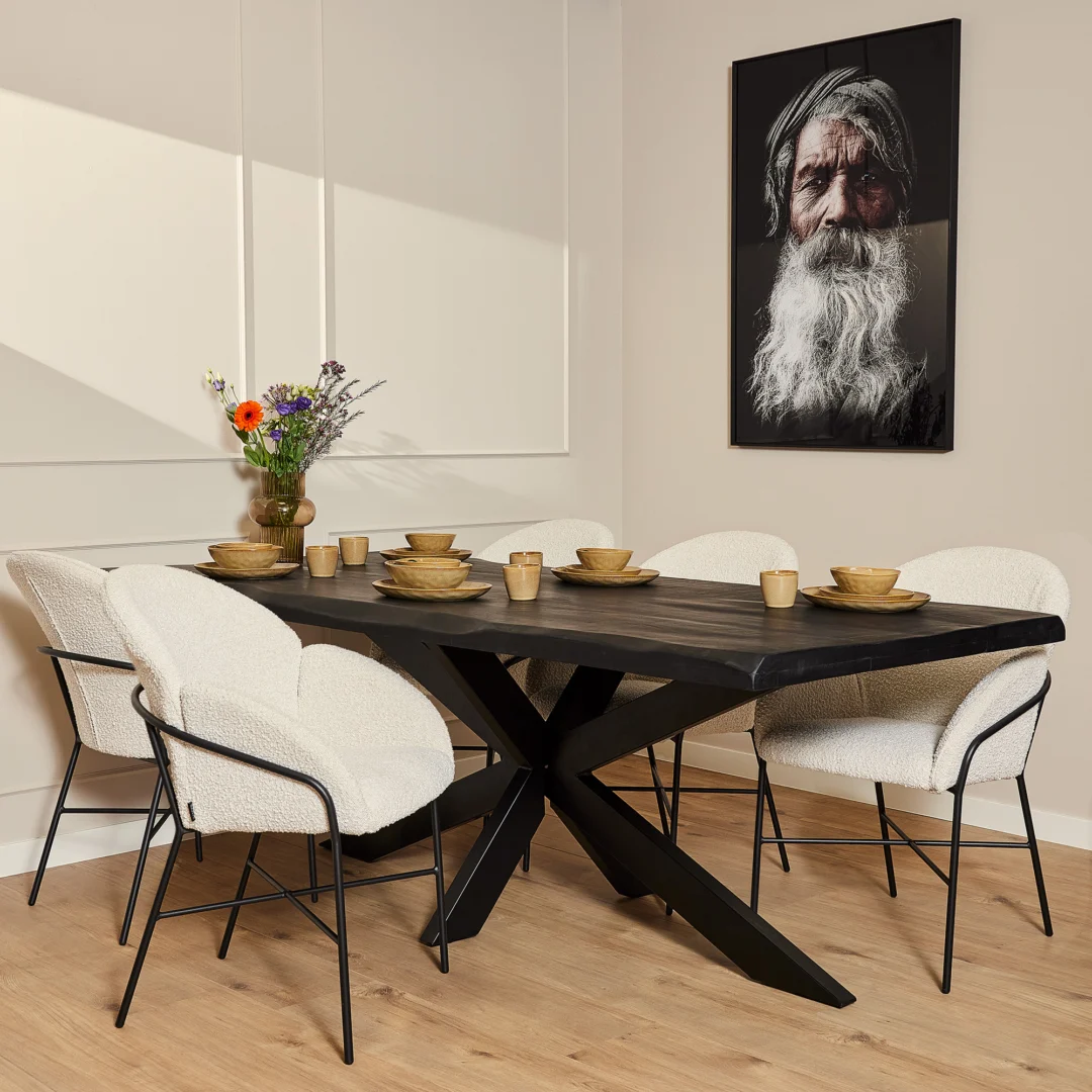 In een moderne eetkamer staat de Boomstamtafel Boston Zwart (220 cm) voor zes personen, omringd door wit gestoffeerde stoelen. Een vaas met bloemen siert de tafel en aan de muur hangt een portret van een oudere man met een lange witte baard.