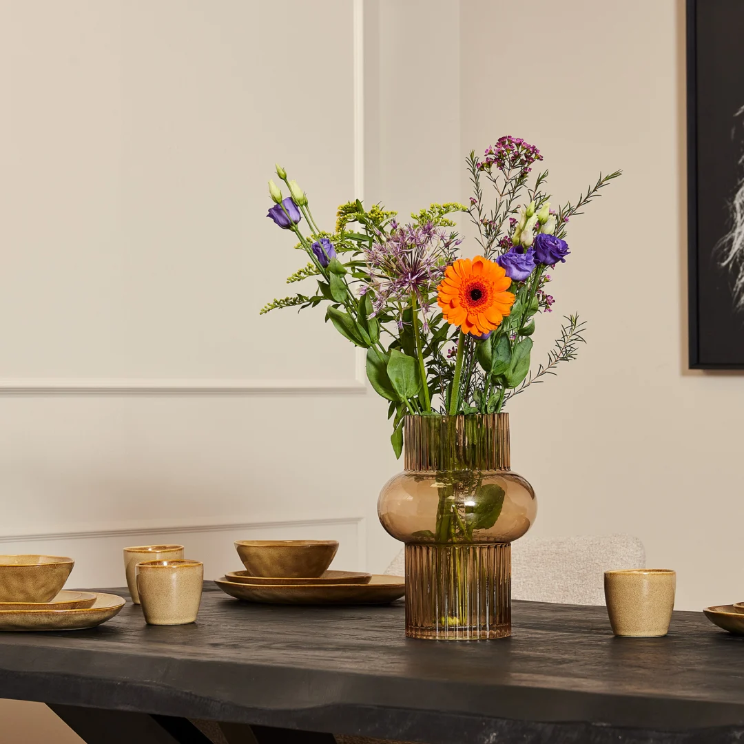 Een bruine glazen vaas met kleurrijke bloemen staat op de Boomstamtafel Boston Zwart (220 cm), bezet met beige keramische kommen, borden en kopjes. Op de achtergrond zie je een lichtgekleurde muur en een glimp van ingelijst kunstwerk.