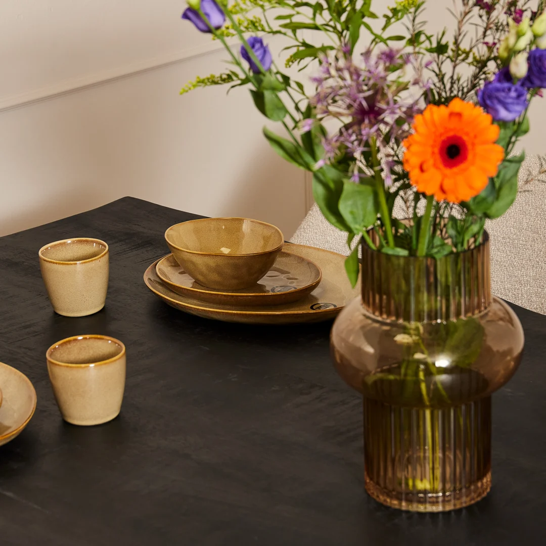 Een Boomstamtafel Boston Zwart (220 cm) set met beige keramische kopjes, kommen, borden en een geribbelde glazen vaas met een levendig boeket paarse en oranje bloemen.