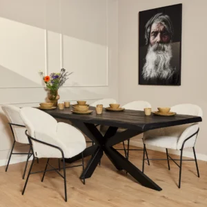 Een moderne eetkamer met de Boomstamtafel Boston Zwart 240 cm, zes witte stoelen met kussens, een bloemenvaas, servies en een groot portret van een oudere man met een lange witte baard aan de muur.