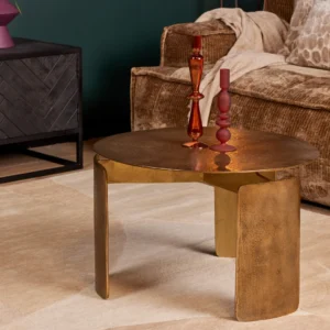 De Salontafel 60 x 60 CM Rond Compact, een ronde goudmetallic salontafel met sculpturale poten, staat op een beige vloerkleed. Bovenop staan een rode en een roze kandelaar. Een bank met bruine textuur en een zwart bijzettafeltje staan op de achtergrond.