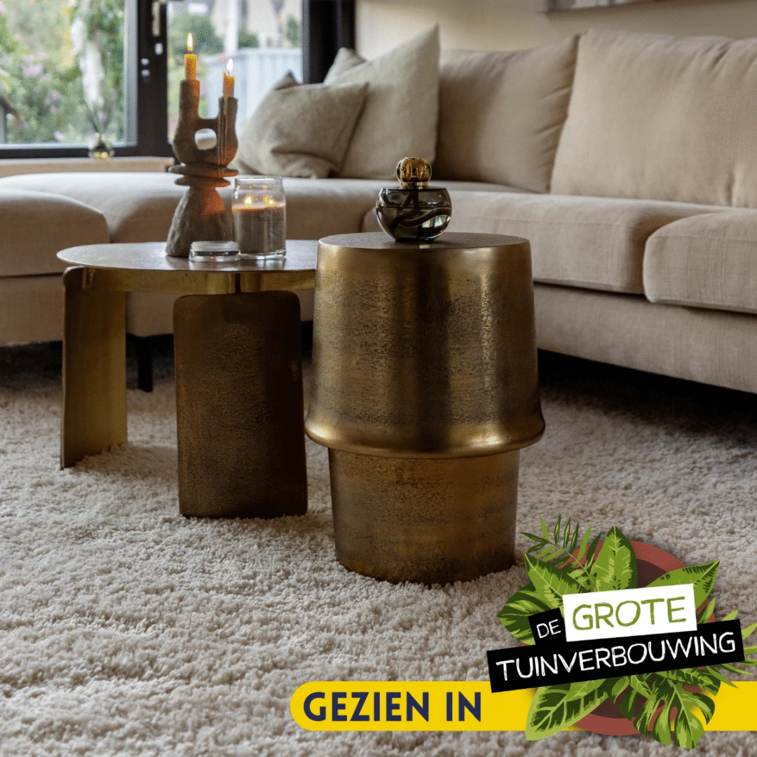 Een woonkamer met een beige sofa, Salontafel 60 x 60 CM Rond Compact en een metalen bijzettafel op een pluche crèmekleurig tapijt. Zonlicht vult de ruimte door de grote ramen. Onder het logo staat "Gezien in De Grote Tuinverbouwing.