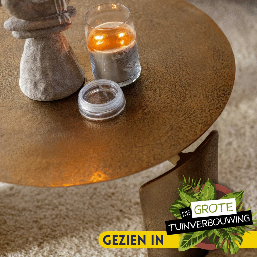 Een Salontafel 60 x 60 CM Rond Compact in brons staat op een beige tapijt, met daarop een brandende kaars in een glazen pot, een open container en een stenen ornament. Het label "GROTE TUINVERBOUWING" staat in de benedenhoek.