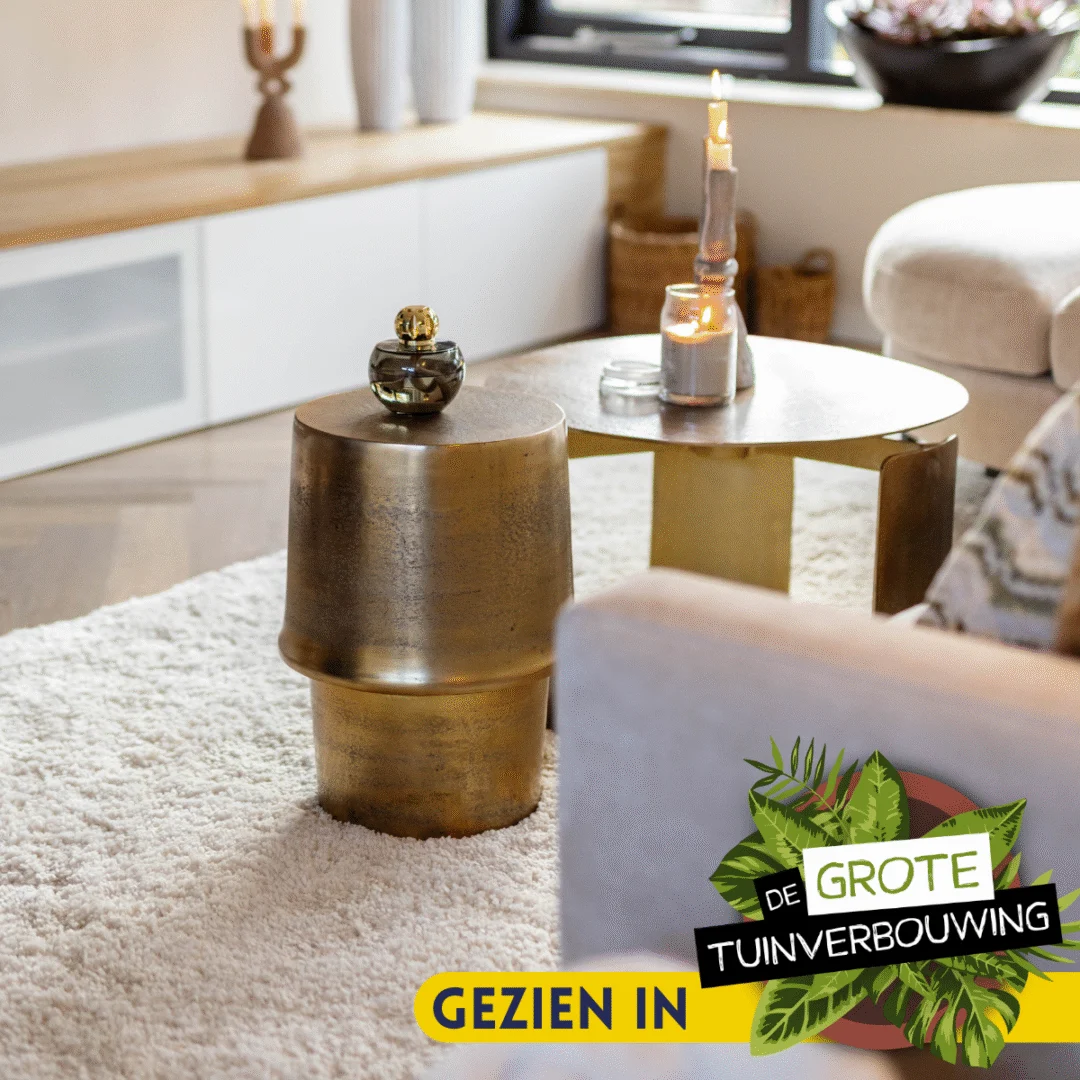 Een gezellige woonkamer met een gouden bijzettafel, de Salontafel 60 x 60 CM Rond Compact, kaarsen en een wit vloerkleed. Een geel "Gezien in De Grote Tuinverbouwing" label verschijnt rechtsonder.