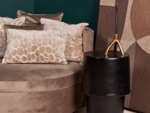 Een beige fluwelen bank met gestructureerde kussens staat bij de bijzettafel Kacy met een decoratieve gouden kandelaar. Een groot abstract kunstwerk leunt tegen een donkergroene muur in deze moderne woonkamer.