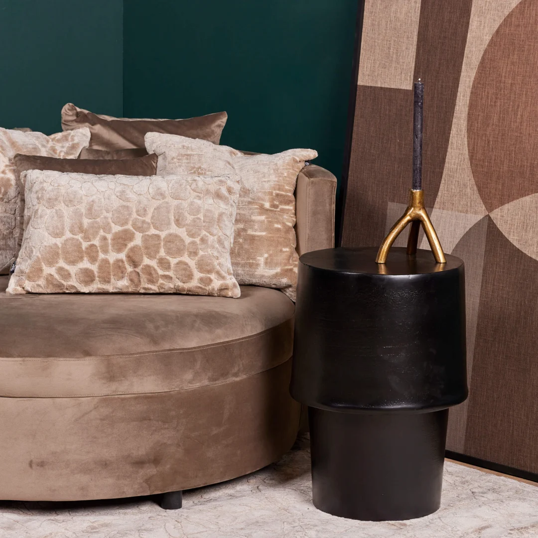 Een beige fluwelen bank met gestructureerde kussens staat bij de bijzettafel Kacy met een decoratieve gouden kandelaar. Een groot abstract kunstwerk leunt tegen een donkergroene muur in deze moderne woonkamer.