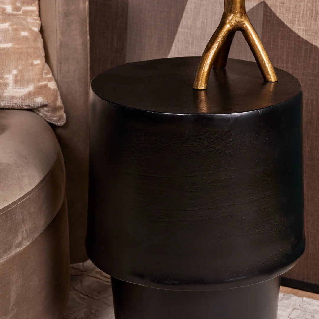 De bijzettafel Kacy, met een modern zwart rond ontwerp, staat naast een beige fluwelen bank. Hierop rust een goudkleurig decoratief object met twee takken, terwijl een beige kussen met patroon op de achtergrond verschijnt.