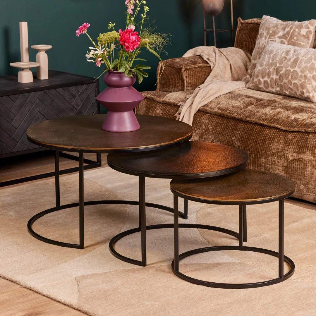 De Salontafel 50 CM Nesting Set, bestaande uit ronde tafels met een donker metalen frame en bruin gestructureerd blad, staat op een beige vloerkleed. Een paarse vaas met kleurrijke bloemen staat op de grootste tafel naast een bruine bank met een beige kleed.