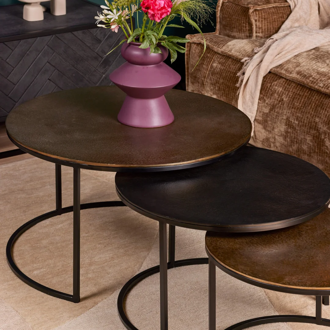 De Salontafel 50 CM Nesting Set bestaat uit drie ronde tafels met donkere metalen poten en tafelbladen in verschillende tinten, met daarop een paarse vaas met roze en witte bloemen, geplaatst naast een bank met bruine textuur en een beige kleed.