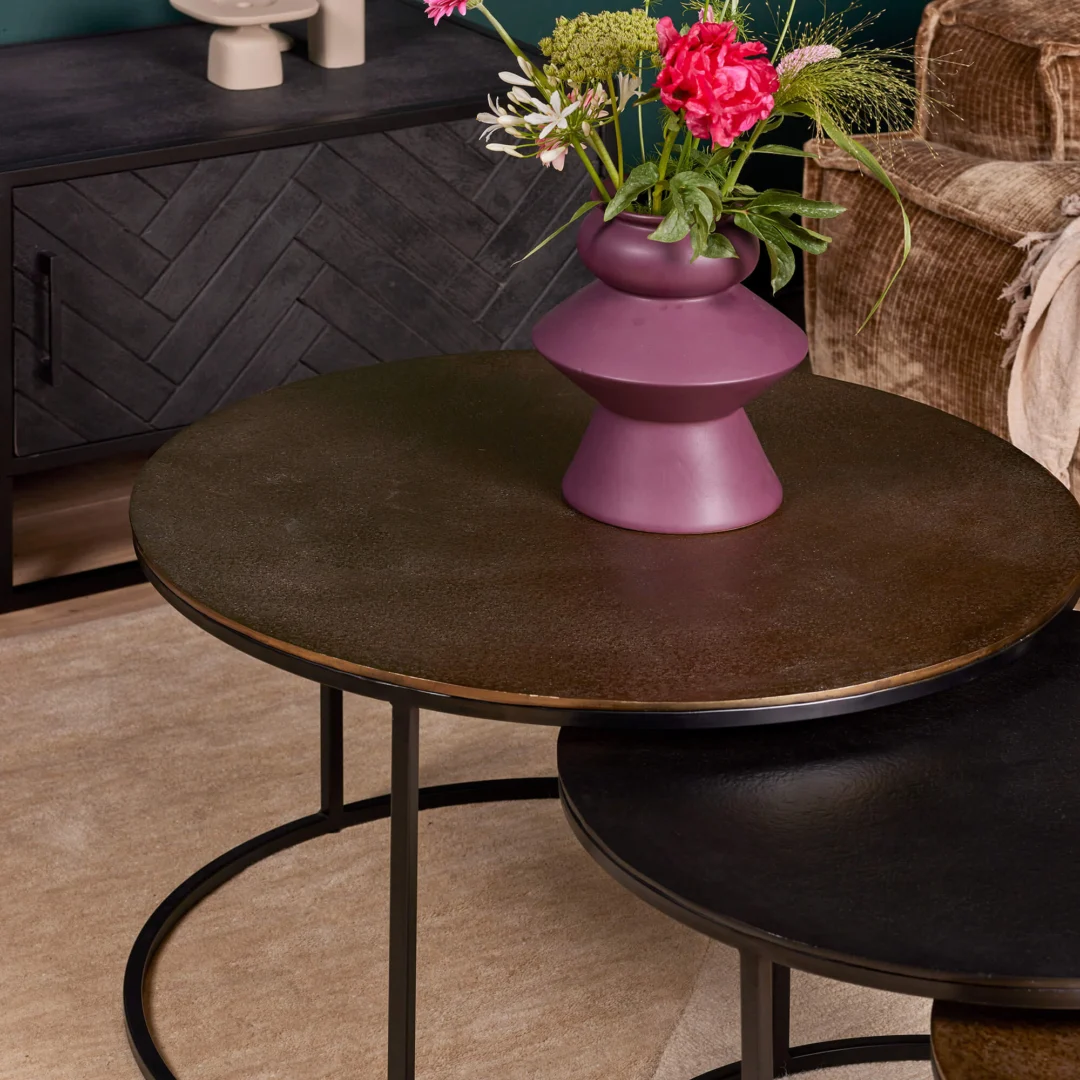 De Salontafel 50 CM Nesting Set, een ronde metalen salontafel met zwarte poten en een donker oppervlak, is vormgegeven met een paarse vaas met roze en groene bloemen. Hij staat naast een kleinere overlappende tafel, op een beige vloerkleed bij een donkere kast.