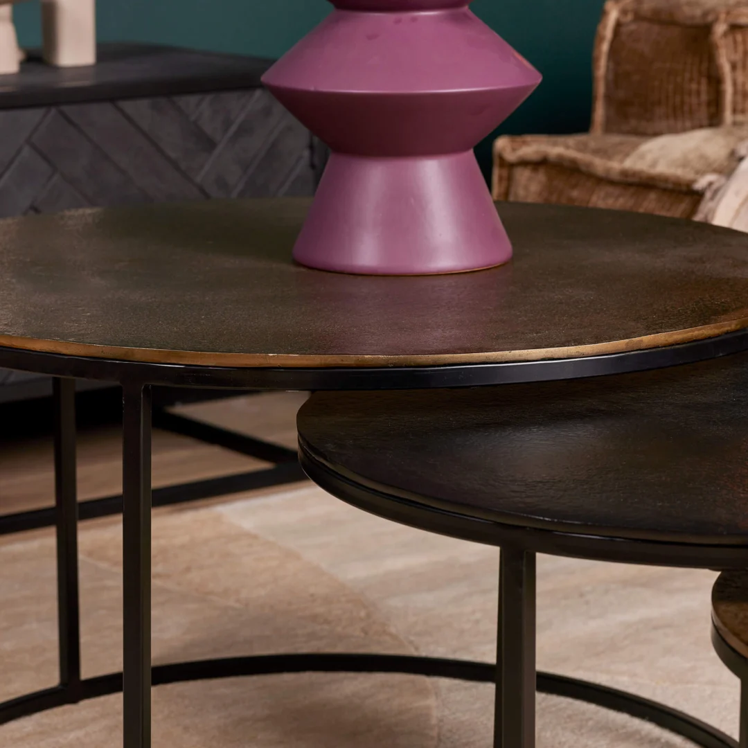 Een close-up van de Salontafel 50 CM Nesting Set - twee ronde zwart metalen tafels met dunne poten. Op de grotere tafel staat een paarse geometrische vaas, terwijl op de achtergrond rieten meubels en een donkere kast zichtbaar zijn.