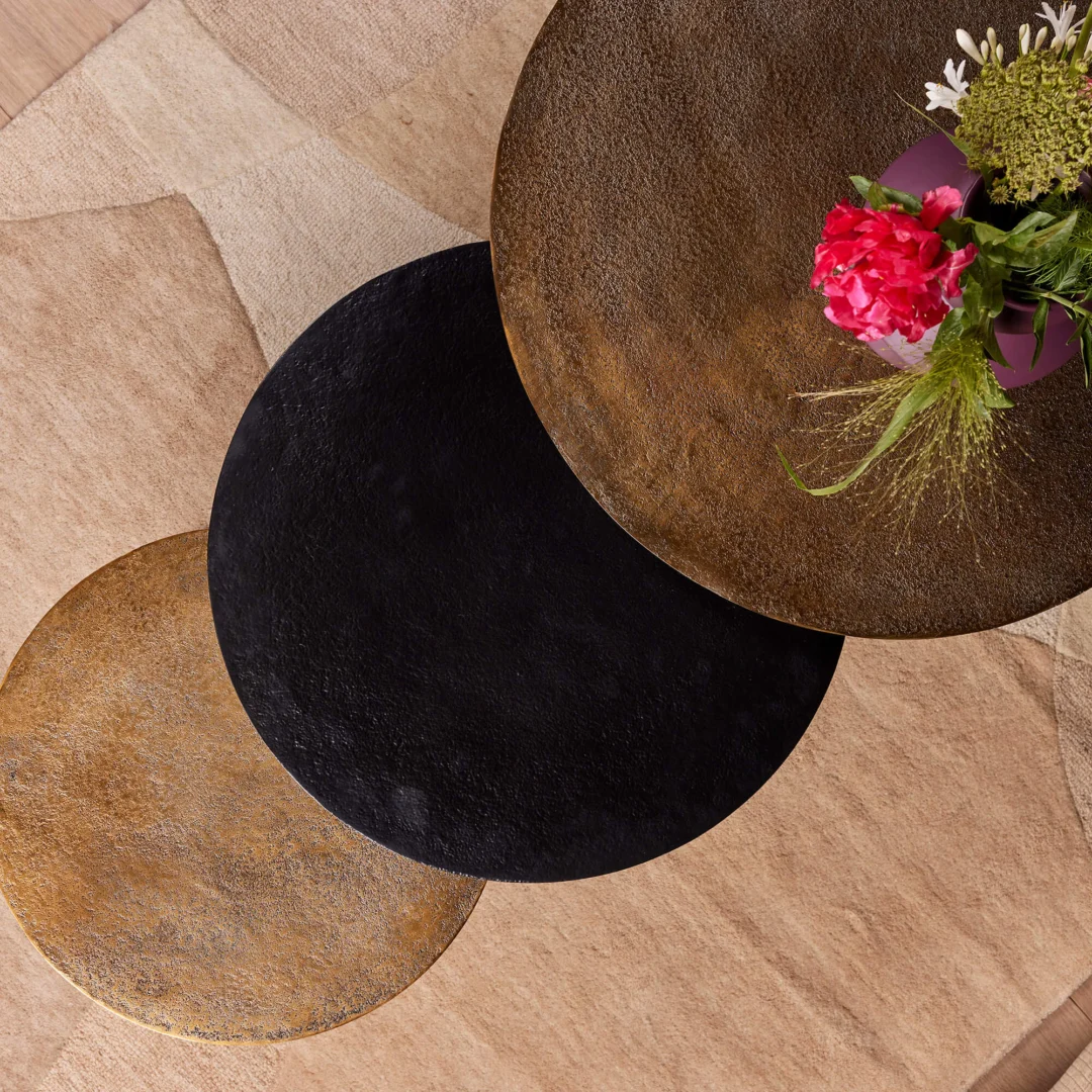 De Salontafel 50 CM Nesting Set, met drie ronde dienbladen in goud, zwart en brons, staat op een beige gestructureerd oppervlak. Op het bovenste dienblad staat een paarse vaas met roze en groene bloemen.