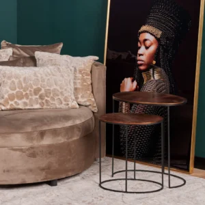 Een ronde beige sofa met gestructureerde kussens staat naast de Bijzettafelset Leaves-two geneste ronde houten tafels. Daarachter hangt een ingelijst portret van een vrouw in koninklijke kleding en gevlochten hoofdtooi tegen een donkergroene muur.