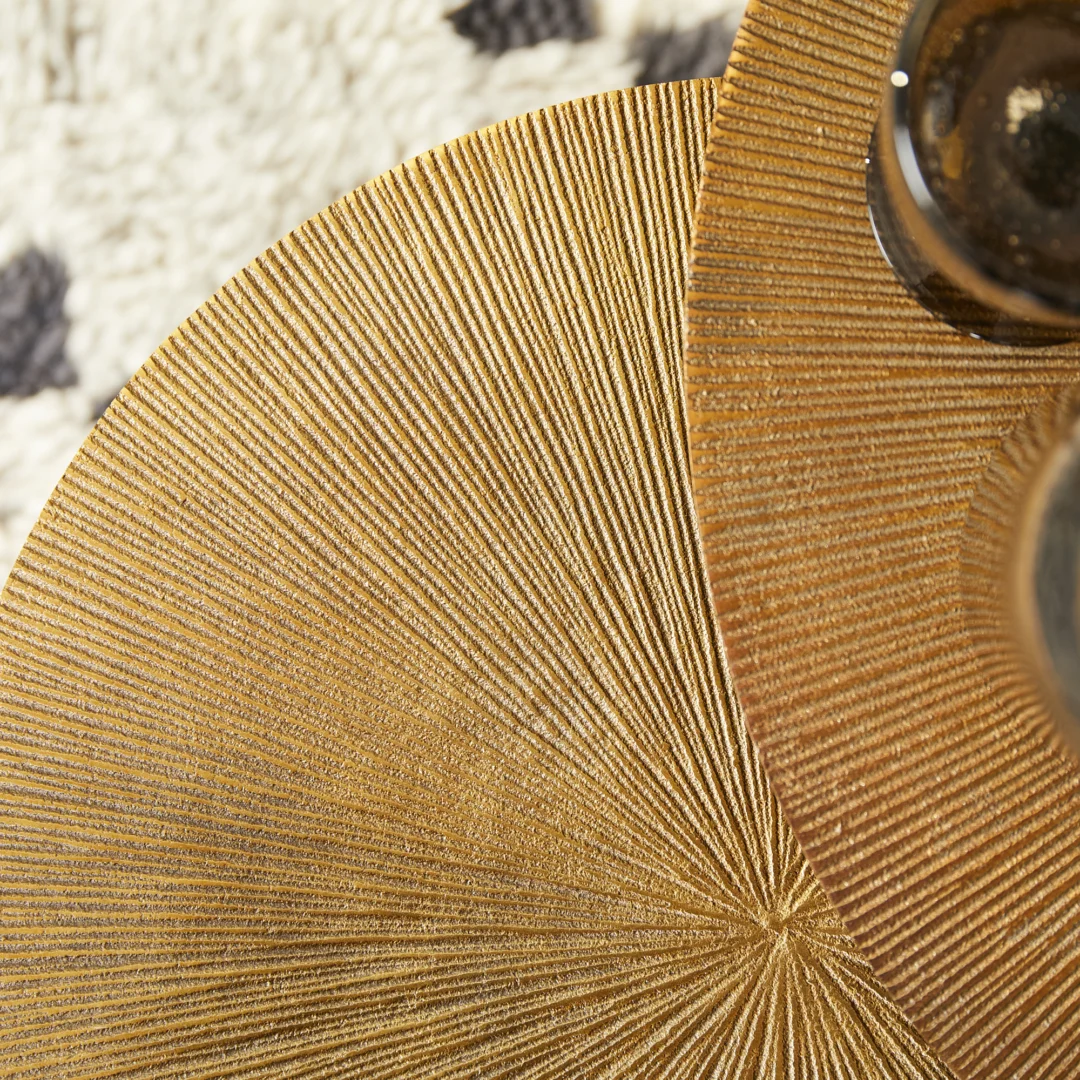 Close-up van twee ronde, gouden Bijzettafels van Ryas met stralende texturen, gedeeltelijk overlappend. Rechtsboven verschijnt een glas met een donker drankje, met op de achtergrond een tapijt met een crèmekleurig en zwart patroon.