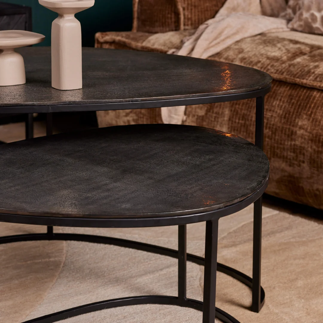 De Salontafel 65 x 50 CM, een ronde zwart metalen salontafel met dunne poten, staat op een lichtbeige vloerkleed in een gezellige woonkamer. Beige keramische vazen sieren de grotere tafel, met een bruine bank met textuur op de achtergrond.