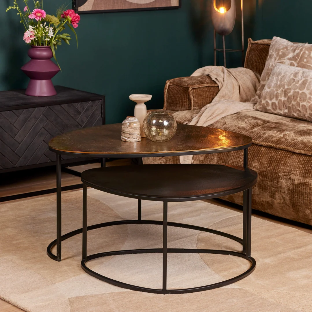 De Salontafel 70 CM Nesting Set, bestaande uit twee ronde salontafels met zwarte metalen frames en donker houten bladen, staat op een beige vloerkleed in een gezellige woonkamer met een bruine bank, zwart dressoir, paarse vaas en donkergroene muren.
