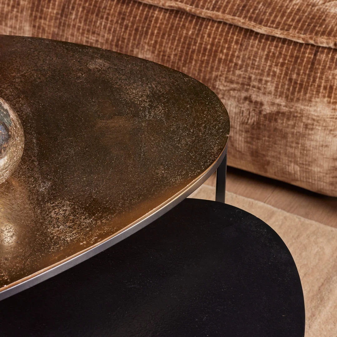 Close-up van de Salontafel 70 CM Nesting Set - een ovale, gestructureerde salontafel met een donker metalen oppervlak - geplaatst voor een pluche bruine ribfluwelen sofa. Een tweede ronde tafel uit de set is gedeeltelijk zichtbaar op de houten vloer.