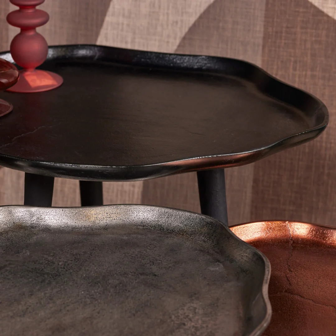 De Bijzettafel set Marna bestaat uit drie metalen dienbladen met golvende randen in de afwerkingen zwart, zilver en koper, op elkaar gestapeld voor een gestructureerde achtergrond. Een rode kandelaarhouder siert de zwarte tafel.