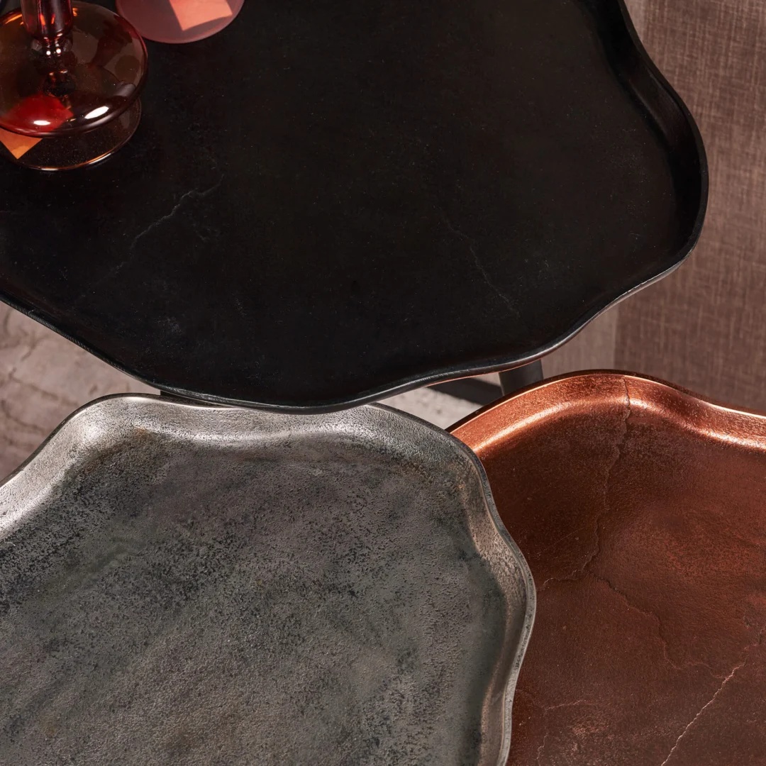 De Bijzettafel set Marna bestaat uit drie metalen dienbladen met golvende randen - zwart, zilver en koperkleurig - met een matte afwerking. Een rood glazen object rust gedeeltelijk op het zwarte dienblad.