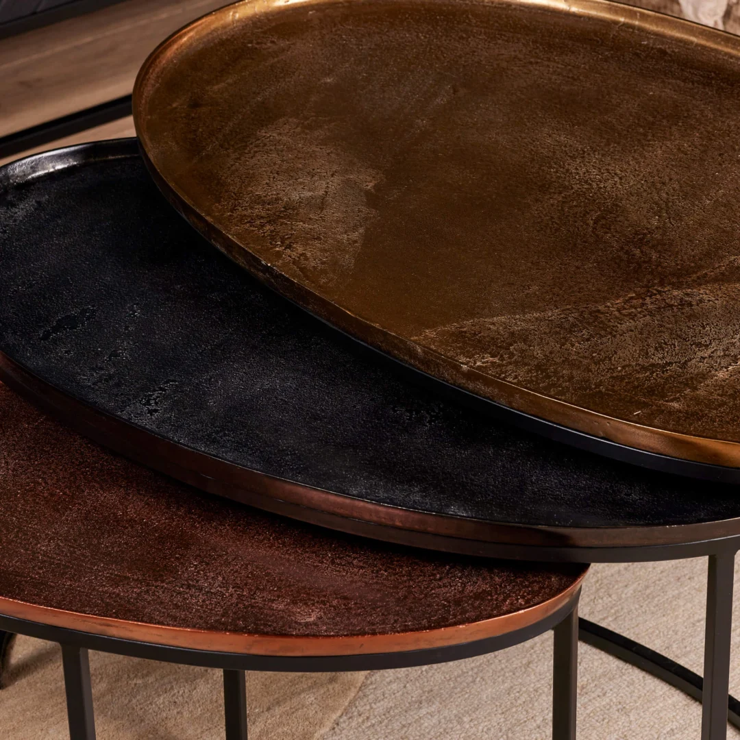 De Salontafel 100 x 60 x 50 CM heeft drie overlappende ovale metalen tafelbladen in zwart, brons en koper met gestructureerde oppervlakken, slanke randen en slanke zwarte poten, gerangschikt in een nestelend patroon.