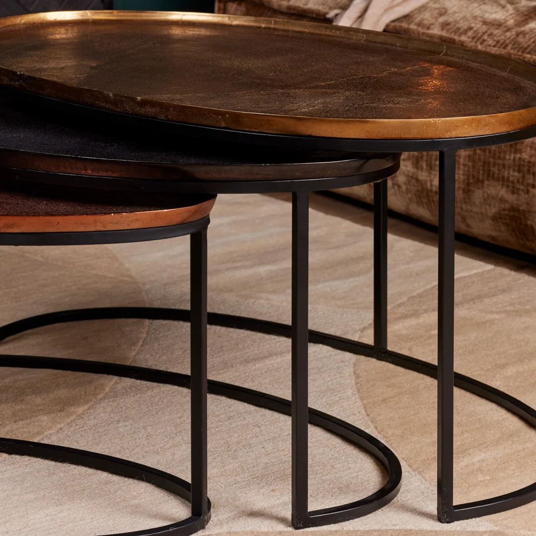 De Salontafel 100 x 60 x 50 CM, een moderne nesttafel met een bronsachtig metalen blad en slanke zwarte poten, staat op een beige vloerkleed en is gedeeltelijk weggestopt onder een tan bank.