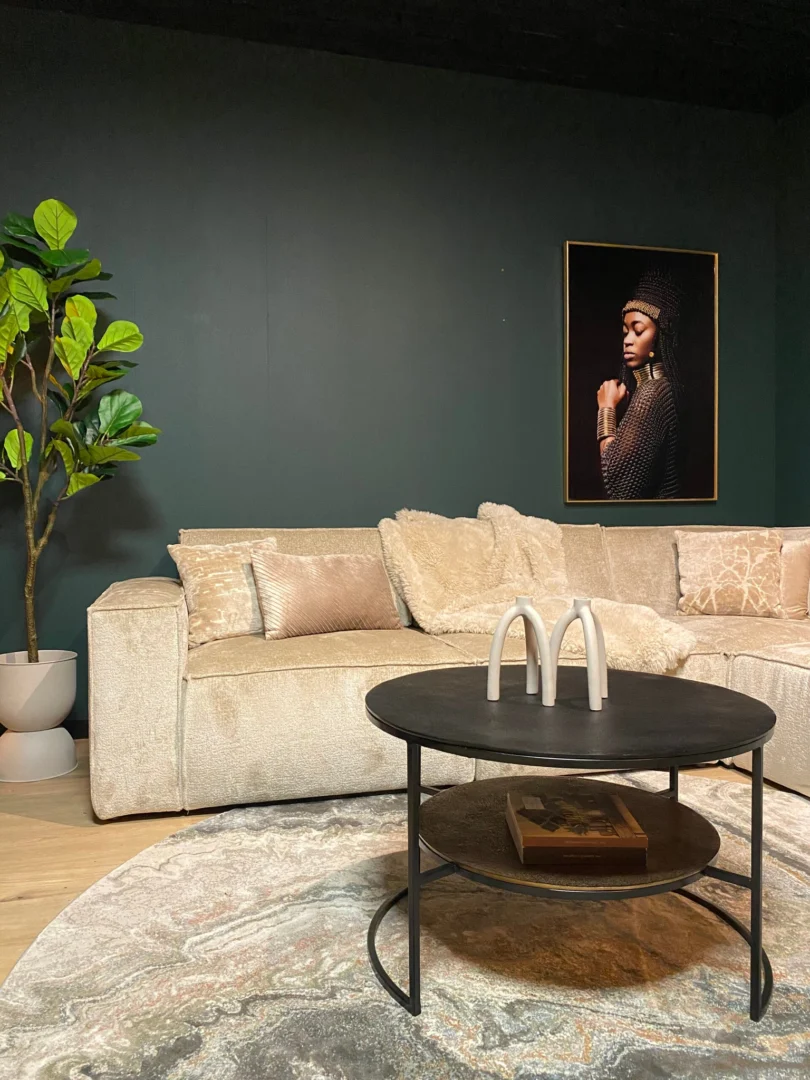 Een moderne woonkamer met een lichtbeige sofa, kussens met structuur, een vierkante zwart-gouden "Salontafel Zwart, Goud 80 x 80 CM," een grote, bebladerde potplant en muurschilderingen van een vrouw tegen een donkergroene muur.