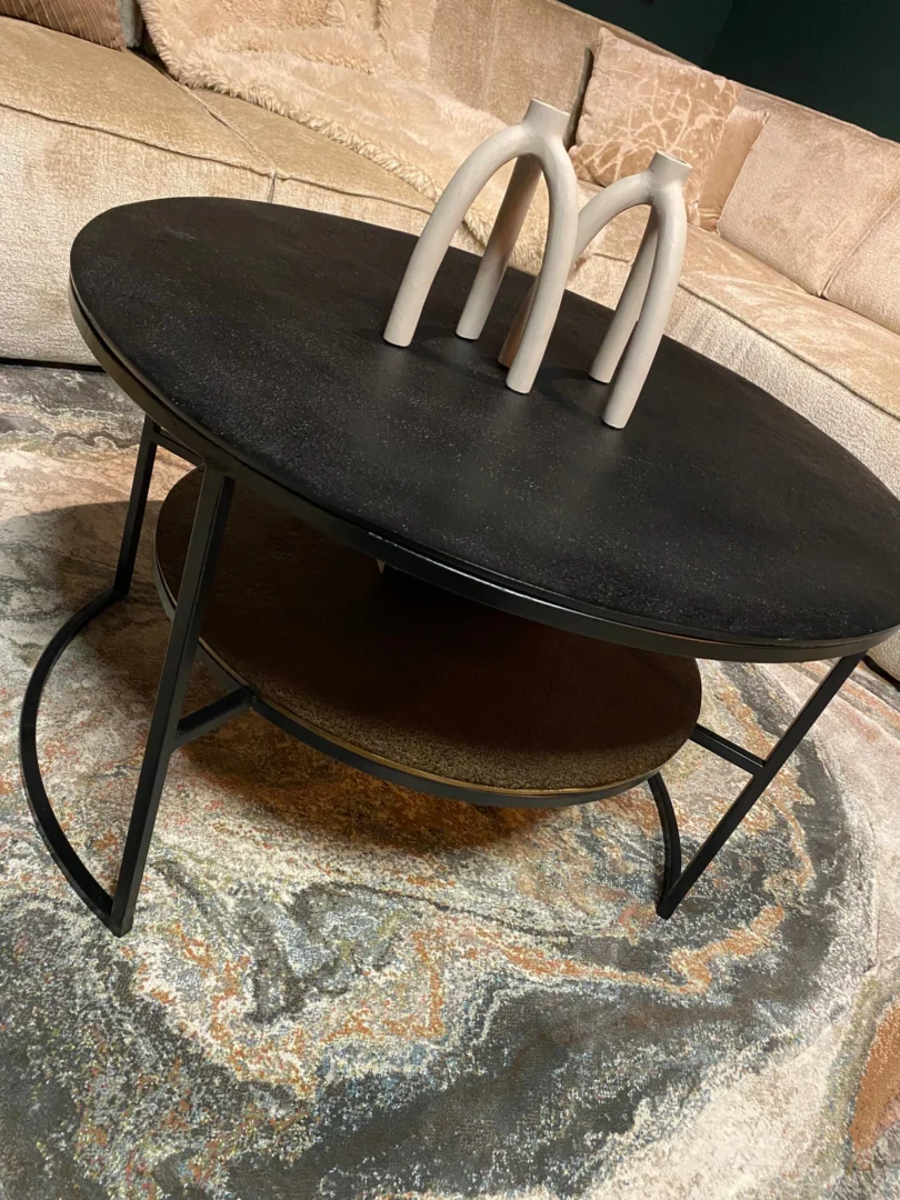 Een vierkante zwart-gouden salontafel (Salontafel Zwart, Goud 80 x 80 CM) met een onderste plank staat op een gedessineerd vloerkleed, met daarop twee gebogen beige keramieken vazen. Op de achtergrond staat een beige sofa.