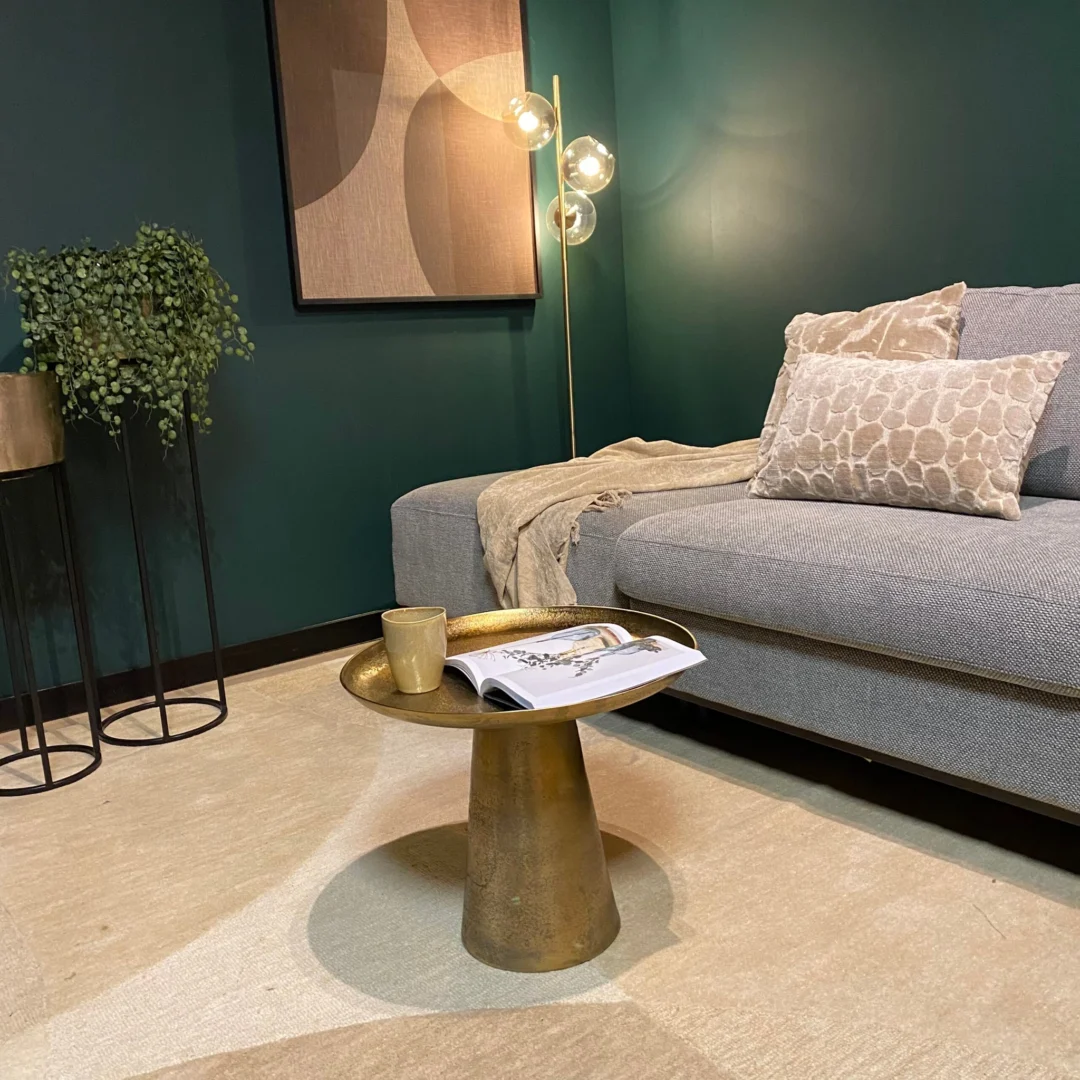 Een moderne woonkamer met een grijze bank, kussens met patroon, een beige kleed, een Salontafel 50 x 50 CM Vierkant met een mok en een tijdschrift, een potplant, abstracte muurkunst en een staande lamp met drie gloeilampen.