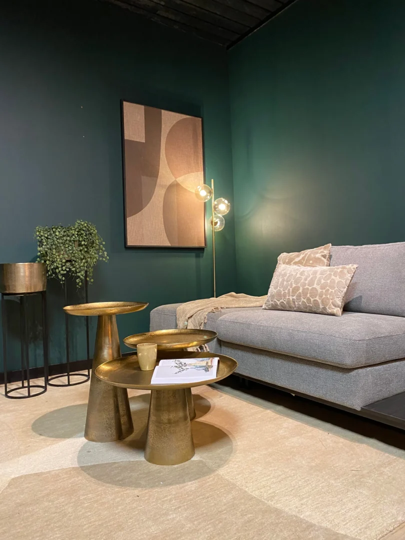 Een moderne woonkamer met donkergroene muren, een grijze bank met gedessineerde kussens, abstracte kunst, een bladplant in een gouden pot en de Salontafel 50 x 50 CM Vierkant als chique accenttafel naast een vloerlamp met drie gloeilampen.