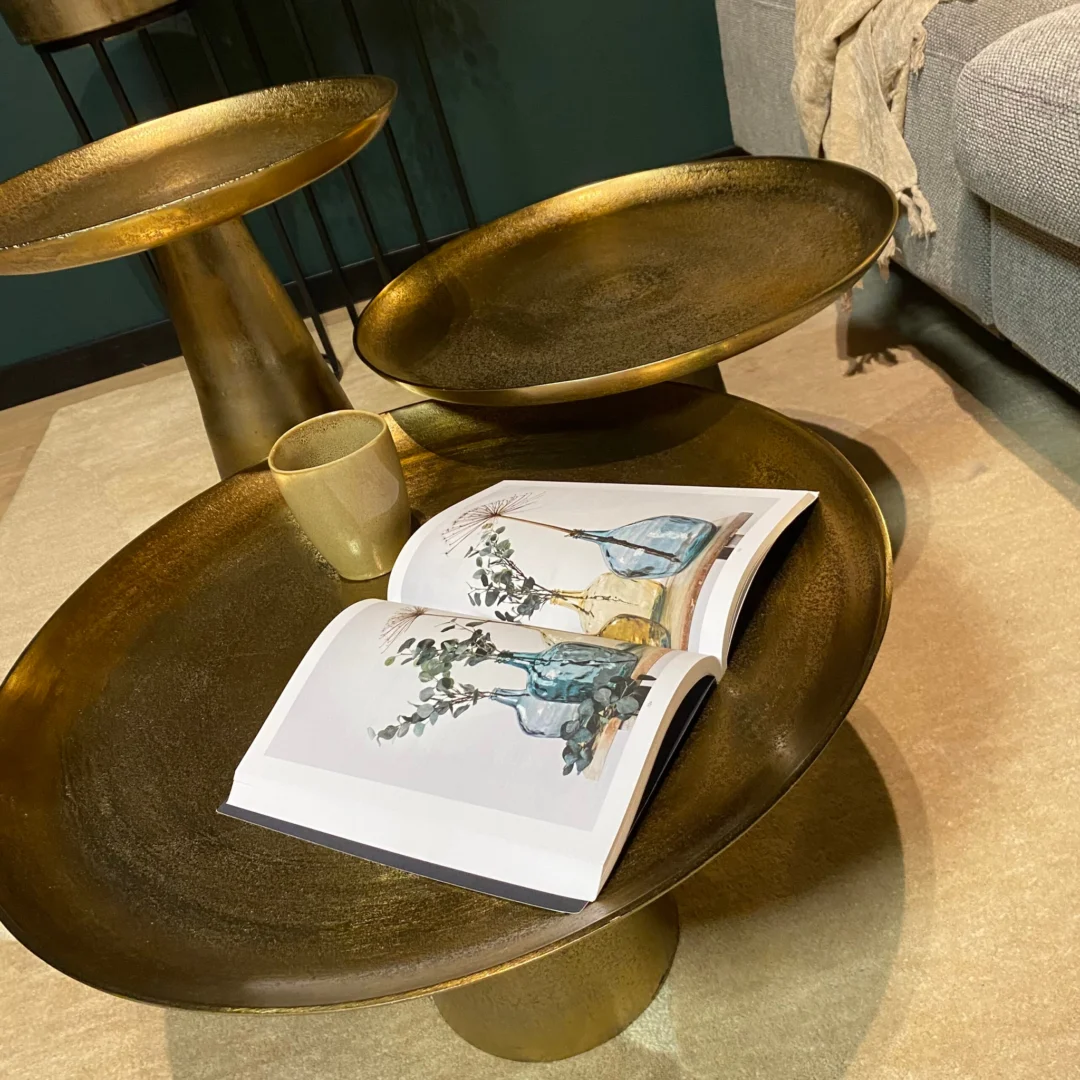 Een moderne woonkamer toont de Salontafel 50 x 50 CM Vierkant. Een opengeslagen kunstboek met plantenillustraties en een gouden beker rusten op de tafel, terwijl een grijze bank met een beige kleed op de achtergrond verschijnt.