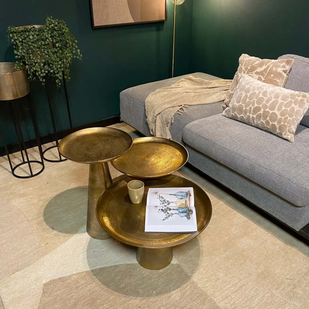 In een moderne woonkamer staat een grijze bank met kussens met een patroon, een beige kleed en de Salontafel 50 x 50 CM Vierkant met een kopje en een tijdschrift. Een plant en lamp staan in de hoek tegen donkergroene muren en een licht vloerkleed.