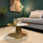 Een moderne woonkamer met een grijze bank, gedessineerde kussens, een pluche beige vloerkleed, een Vierkant Salontafel 58 x 36 CM met een mok en een tijdschrift, een staande lamp met gloeilampen, een hangplant en abstracte muurkunst.
