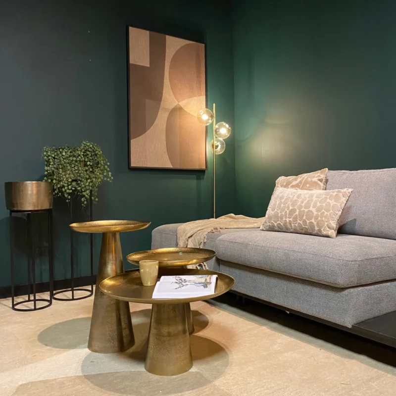 Moderne woonkamer met donkergroene muren, een grijze bank met gedessineerde kussens, gouden accenttafels zoals de Salontafel 58 x 36 CM Vierkant, een plant op een standaard, abstracte muurkunst en een vloerlamp met gloeilamp voor een gezellige, stijlvolle sfeer.