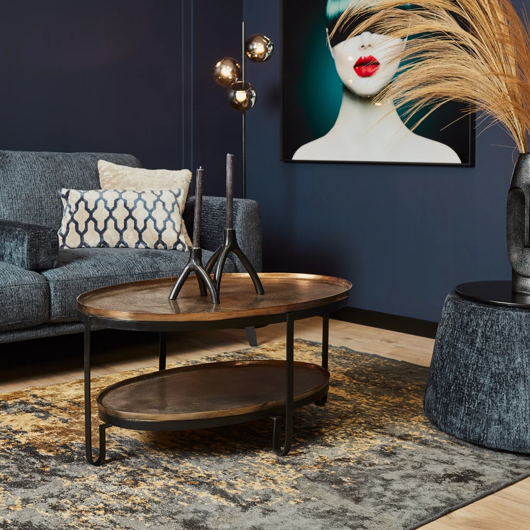 Een moderne woonkamer met een blauwe bank, decoratieve kussens, een abstract vloerkleed en een eigentijdse inrichting. Het middelpunt is de Salontafel 98 CM Ovaal salontafel. Een gewaagd kunstwerk van een vrouw met rode lippen accentueert de donkerblauwe muur.