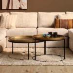 Twee ronde salontafels, goudkleurig blad met zwarte rand en poten. Zachte, lichte sofa en kleed op de achtergrond. Warm, natuurlijk licht.