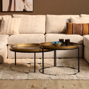 Twee ronde salontafels, goudkleurig blad met zwarte rand en poten. Zachte, lichte sofa en kleed op de achtergrond. Warm, natuurlijk licht.