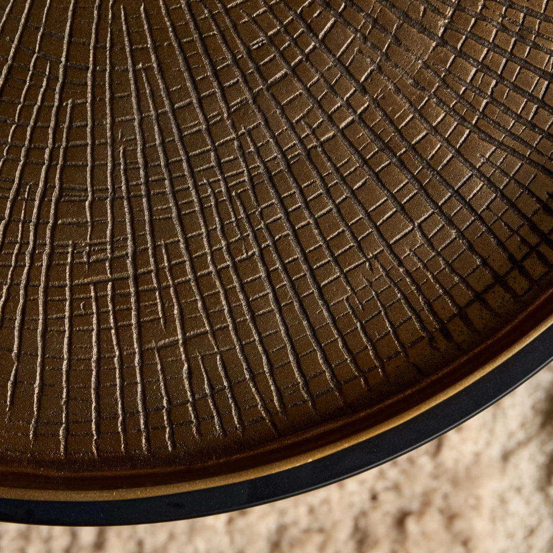 Detailopname van een bruine, ronde salontafel met een patroon van lijnen. Het tafelblad heeft een zwarte rand. Zachte belichting.