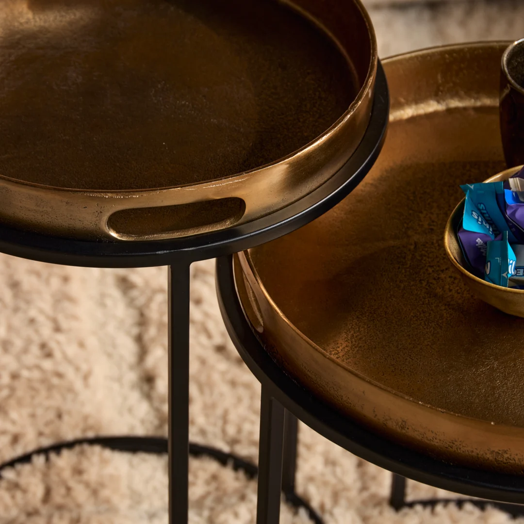 Close-up van een gouden design bijzettafel met zwarte poten. De tafels hebben een dienbladachtig oppervlak en staan op een zacht, wit tapijt.