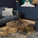 Een moderne woonkamer heeft een donkergrijze bank, kussen met patroon, Salontafel 80 x 80 CM Vierkant Design salontafel met twee zwarte kandelaars, abstract vloerkleed, minimalistisch kunstwerk en staande lamp tegen donkerblauwe muren.