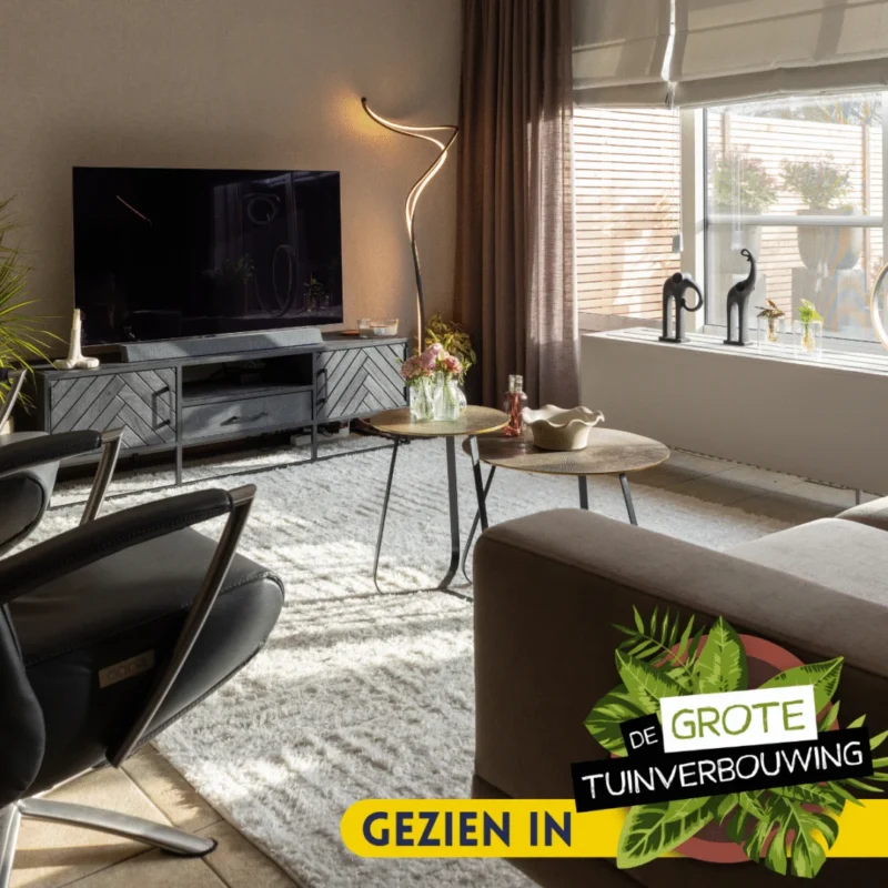 Een moderne woonkamer heeft een tv, zwarte fauteuil, bruine bank, Salontafel 40 x 50 CM Vierkant als salontafel en een groot zonovergoten raam. Op tafel staan decoratieve planten en drankjes. Tekst "GEZIEN IN DE GROTE TUINVERBOUWING" is overlaid.