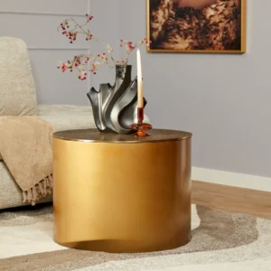 De Salontafel Fiore Goud, een ronde gouden salontafel, toont een moderne zwarte vaas met rode bessen en een witte kaars in een amberkleurige houder. Een beige bank, abstracte muurkunst en een zacht vloerkleed maken deze gezellige woonkamer compleet.