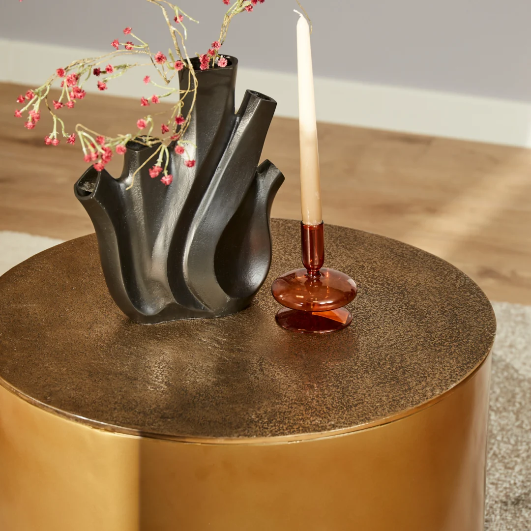 De Salontafel Fiore Goud, een ronde goudkleurige tafel, is binnenshuis gestyled met een zwarte vaas met roze bloemen, een hoge witte kaars in een rode houder, een houten vloer en een neutraal vloerkleed.