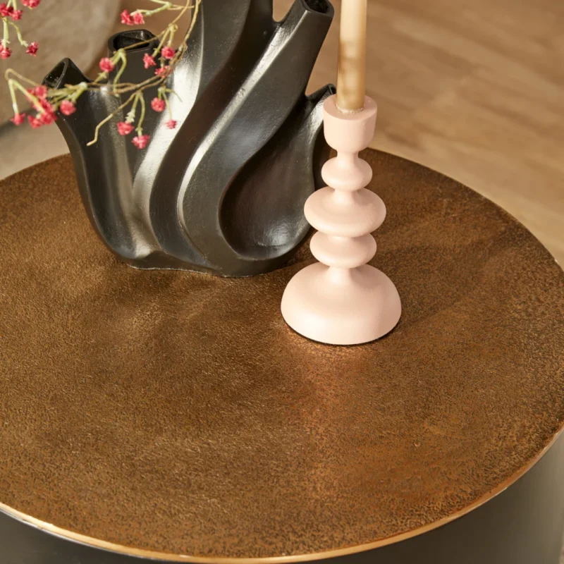 Een ronde, gestructureerde Salontafel Fiore | Zwart/Goud toont een sculpturale zwarte vaas met roze bloemen en een lichtroze kandelaar met een beige kaars.