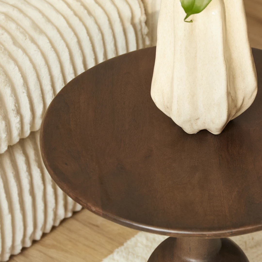De bijzettafel Donna, rond en donker hout, houdt een crèmekleurige gebeeldhouwde vaas van keramiek met een groen blad naast een beige ribfluwelen bank en een licht vloerkleed op een houten vloer.