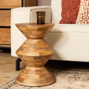 Bijzettafel Abel, een klein geometrisch houten tafeltje, staat naast een lichtgekleurde bank met gestructureerde kussens en een glazen beker bovenop, geplaatst op een vloerkleed met patroon in een gezellige woonkamer.