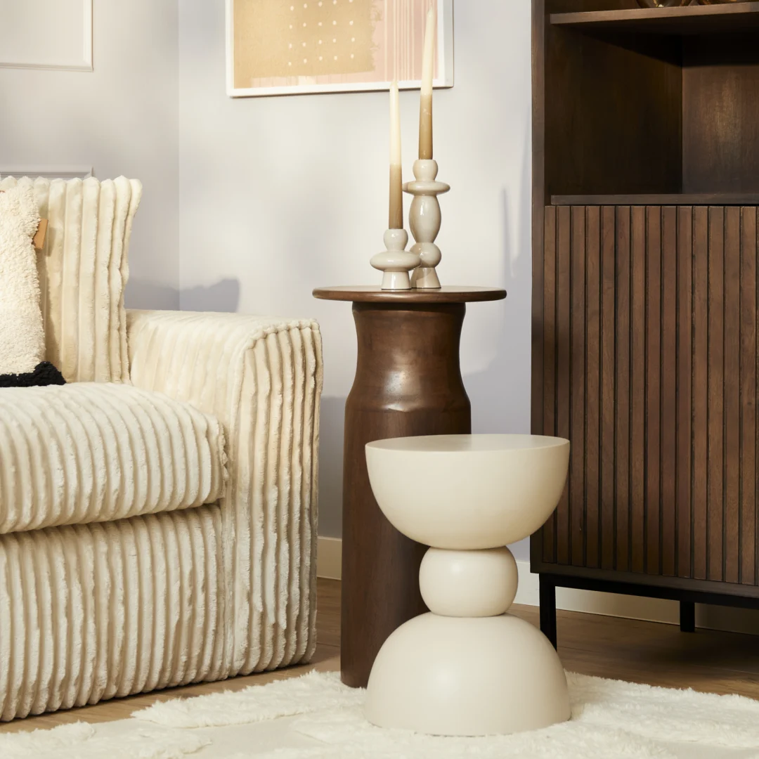 Een gezellige woonkamerhoek met een crèmekleurige ribfluwelen bank, de ronde donkerhouten bijzettafel van Mason met sculpturale beige kandelaars en een moderne gebroken witte sculpturale kruk op een licht vloerkleed naast een houten kast.