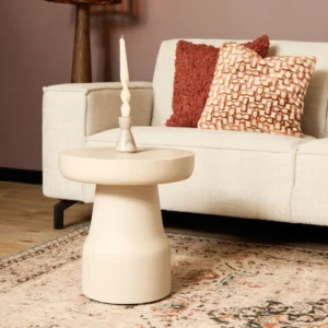 Een moderne woonkamer met een beige bank, twee gedessineerde kussens, een bijzettafel met een crèmekleurige gedraaide kaars en een gedessineerd tapijt op een houten vloer. De muur is mauve geschilderd.