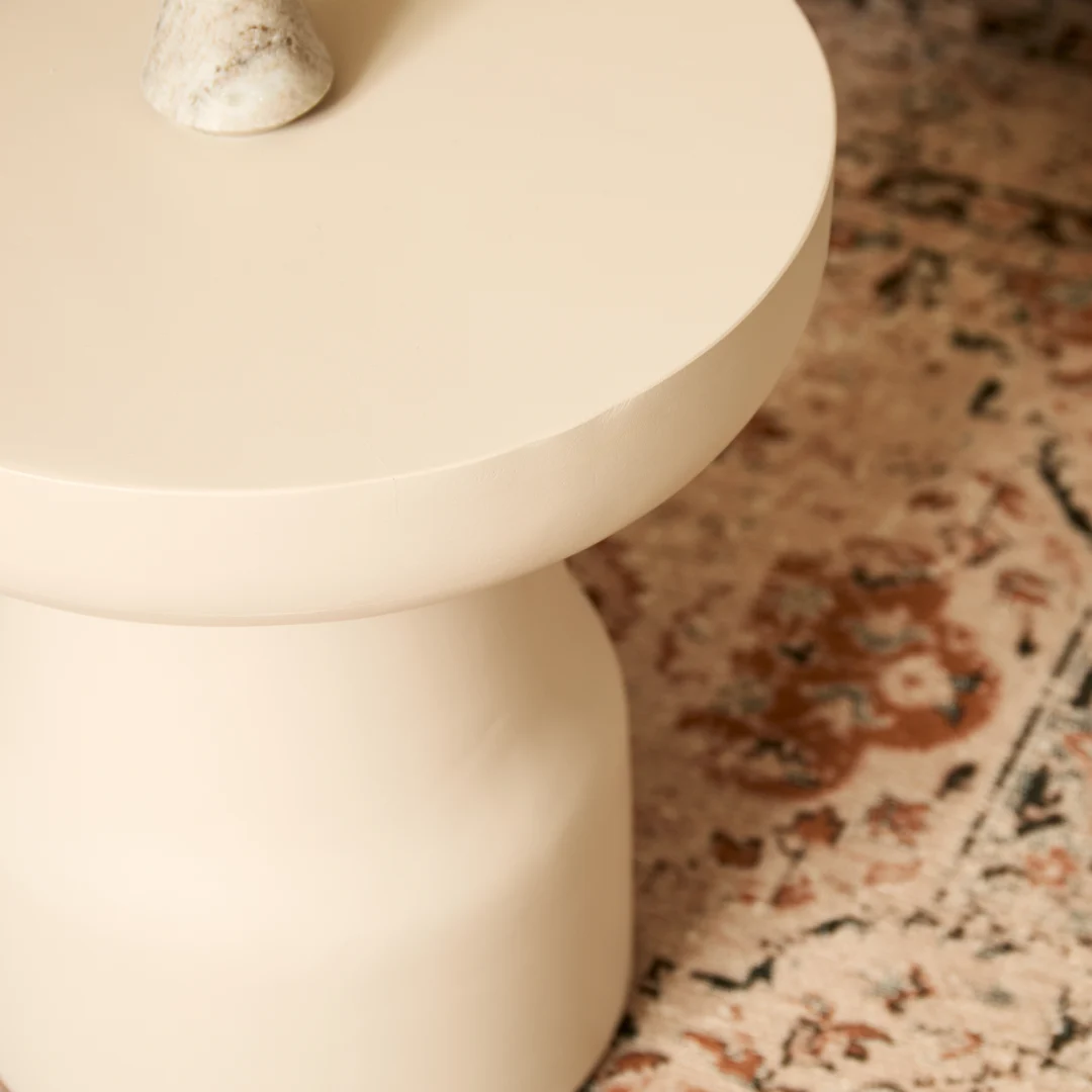 De bijzettafel Drum, een modern rond stuk in crème met een sculpturale basis, staat op een gedessineerd tapijt in beige, roze en bruine tinten, met daarop een klein lichtgekleurd object.