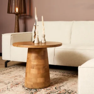 De ronde houten bijzettafel Timo heeft drie sculpturale kandelaars en staat op een gedessineerd tapijt tussen twee crèmekleurige banken in een gezellige, zacht verlichte woonkamer. Een hoge staande lamp staat in de hoek.